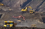 Heavy machinery operates on a large-scale mining site. Excavators and dump trucks are moving earth and materials, surrounded by rocky terrain and steep slopes. The equipment is primarily yellow and orange, indicating industrial use, and work is being actively carried out to extract resources.