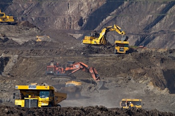 Heavy machinery operates on a large-scale mining site. Excavators and dump trucks are moving earth and materials, surrounded by rocky terrain and steep slopes. The equipment is primarily yellow and orange, indicating industrial use, and work is being actively carried out to extract resources.