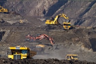 Heavy machinery operates on a large-scale mining site. Excavators and dump trucks are moving earth and materials, surrounded by rocky terrain and steep slopes. The equipment is primarily yellow and orange, indicating industrial use, and work is being actively carried out to extract resources.