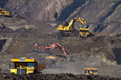 Heavy machinery operates on a large-scale mining site. Excavators and dump trucks are moving earth and materials, surrounded by rocky terrain and steep slopes. The equipment is primarily yellow and orange, indicating industrial use, and work is being actively carried out to extract resources.