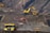 Heavy machinery operates on a large-scale mining site. Excavators and dump trucks are moving earth and materials, surrounded by rocky terrain and steep slopes. The equipment is primarily yellow and orange, indicating industrial use, and work is being actively carried out to extract resources.