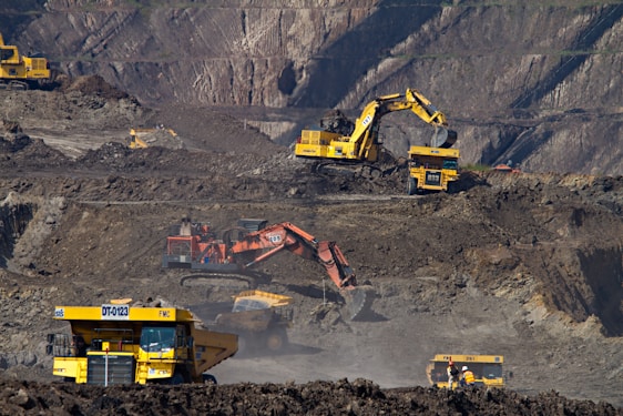 Heavy machinery operates on a large-scale mining site. Excavators and dump trucks are moving earth and materials, surrounded by rocky terrain and steep slopes. The equipment is primarily yellow and orange, indicating industrial use, and work is being actively carried out to extract resources.
