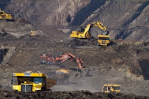 Heavy machinery operates on a large-scale mining site. Excavators and dump trucks are moving earth and materials, surrounded by rocky terrain and steep slopes. The equipment is primarily yellow and orange, indicating industrial use, and work is being actively carried out to extract resources.