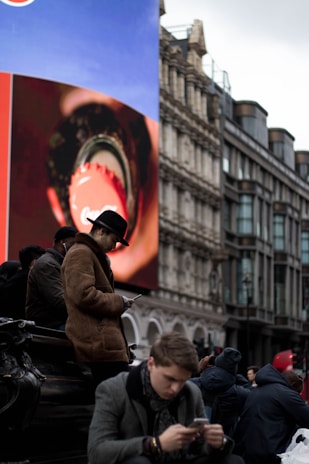 A bustling urban scene features several people sitting and using their phones beneath a large digital billboard displaying a close-up advertisement. The background includes classic architectural buildings, adding a contrast between the historic and modern elements.