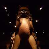 A large wooden totem or sculpture stands prominently against a dark background, illuminated by soft lighting. The figure has an animal-like face with prominent features and raised hands, each holding a smaller carved face.