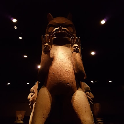 A large wooden totem or sculpture stands prominently against a dark background, illuminated by soft lighting. The figure has an animal-like face with prominent features and raised hands, each holding a smaller carved face.