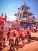 A group of people, predominantly young men, are carrying a large wooden structure adorned with colorful decorations in front of a traditional, ornate temple with a tiered roof. The setting appears to be during a festive occasion, as the participants are engaged in a communal activity under a clear blue sky.