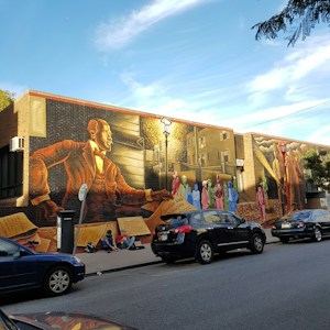 A large mural on the side of a brick building features a historical figure in a suit reaching out with a scroll, surrounded by colorful, diverse figures. The scene includes architectural elements like windows, a streetlamp, and a staircase, with people sitting on the sidewalk nearby. Several cars are parked along the street.