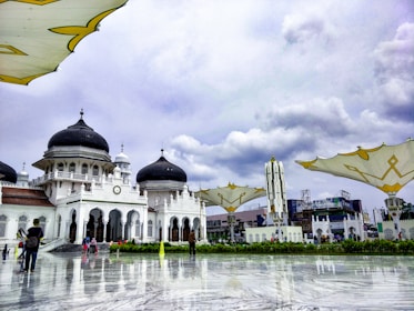 An elegant mosque with striking black domes and intricate white arches surrounded by a large open courtyard. Modern structures with large umbrella-like shades are visible in the foreground. A few people are scattered throughout the area, some walking, some taking photos. The sky is overcast, casting a grayscale hue over the scenery.