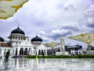 An elegant mosque with striking black domes and intricate white arches surrounded by a large open courtyard. Modern structures with large umbrella-like shades are visible in the foreground. A few people are scattered throughout the area, some walking, some taking photos. The sky is overcast, casting a grayscale hue over the scenery.