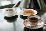 A cup of tea in a white teacup is placed on a saucer next to a plate holding a pastry. Another ornate cup filled with a dark beverage sits in the foreground, on an intricately designed saucer. The setting is on a glossy table surface, possibly in a cozy café environment.