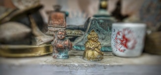 A collection of small, intricately detailed figures and objects placed side by side, including two bronze and gold-tone figurines in traditional attire standing in front of a white cup with a red floral design. The background is blurred, focusing attention on the central objects.