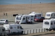 Several caravans are parked near a beach, with a few people walking and a parked red car next to one of the caravans. The beach and ocean are visible in the background, along with a paved path beside the sand.