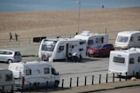 Several caravans are parked near a beach, with a few people walking and a parked red car next to one of the caravans. The beach and ocean are visible in the background, along with a paved path beside the sand.