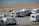 Several caravans are parked near a beach, with a few people walking and a parked red car next to one of the caravans. The beach and ocean are visible in the background, along with a paved path beside the sand.
