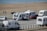 Several caravans are parked near a beach, with a few people walking and a parked red car next to one of the caravans. The beach and ocean are visible in the background, along with a paved path beside the sand.