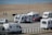 Several caravans are parked near a beach, with a few people walking and a parked red car next to one of the caravans. The beach and ocean are visible in the background, along with a paved path beside the sand.