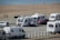 Several caravans are parked near a beach, with a few people walking and a parked red car next to one of the caravans. The beach and ocean are visible in the background, along with a paved path beside the sand.