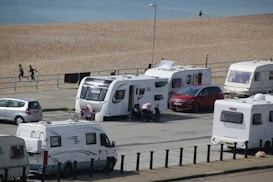 Several caravans are parked near a beach, with a few people walking and a parked red car next to one of the caravans. The beach and ocean are visible in the background, along with a paved path beside the sand.