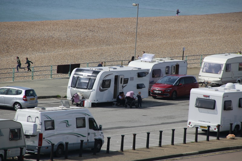 Several caravans are parked near a beach, with a few people walking and a parked red car next to one of the caravans. The beach and ocean are visible in the background, along with a paved path beside the sand.
