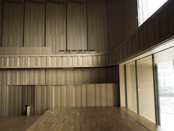 Spacious interior with wooden paneling covering the walls and floors, creating a minimalist and modern aesthetic. Large windows on the right side allow natural light to brighten the space. The design is sleek with clean lines, and there are several chairs positioned in an upper section.
