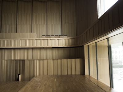 Spacious interior with wooden paneling covering the walls and floors, creating a minimalist and modern aesthetic. Large windows on the right side allow natural light to brighten the space. The design is sleek with clean lines, and there are several chairs positioned in an upper section.
