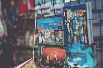 A collection of postcards displayed in a retail stand, featuring images of landmarks and attractions from Munich. The postcards are colorful and include architectural and scenic views.