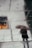 Wet tiles reflect the surrounding environment, showing the faint silhouette of an individual holding an umbrella. There are subtle footprints on the tiles, and a warm glow of light is visible on the left side, contrasting with the muted tones of the pavement.