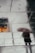 Wet tiles reflect the surrounding environment, showing the faint silhouette of an individual holding an umbrella. There are subtle footprints on the tiles, and a warm glow of light is visible on the left side, contrasting with the muted tones of the pavement.