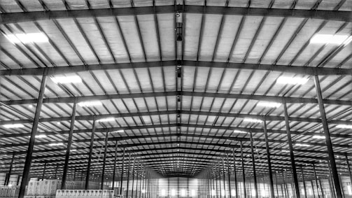 The image features the interior of a large warehouse with a high ceiling, supported by numerous evenly spaced beams and columns. The uniform pattern of lights on the ceiling creates a striking visual effect. Rows of tall, rectangular shelves or boxes are visible at the sides, suggesting storage facilities.