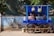 A blue street cart with large wheels displays hanging lemons and various bottled beverages, including soft drinks. A woman is standing behind the cart, partially visible. There is a clay pot on the cart, and a mural of a temple is painted on the wall behind it.