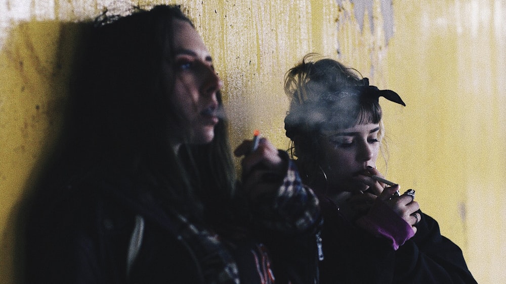 two women smoking while leaning on yellow wall