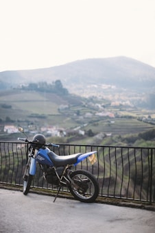 A blue motorcycle is parked against a metal railing overlooking a scenic landscape with rolling hills and a village in the background.