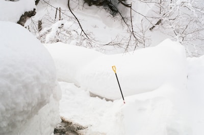 A snowy landscape with large mounds of snow surrounding a narrow path. A single shovel with a yellow handle is stuck in the snow, adding a pop of color. Bare tree branches are visible against an overcast sky.