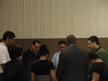 A group of people standing in a circle, appearing to be engaged in a moment of reflection or prayer. The setting is indoors with a neutral-colored background.