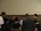 A group of people standing in a circle, appearing to be engaged in a moment of reflection or prayer. The setting is indoors with a neutral-colored background.