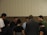 A group of people standing in a circle, appearing to be engaged in a moment of reflection or prayer. The setting is indoors with a neutral-colored background.