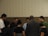 A group of people standing in a circle, appearing to be engaged in a moment of reflection or prayer. The setting is indoors with a neutral-colored background.