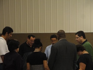 A group of people standing in a circle, appearing to be engaged in a moment of reflection or prayer. The setting is indoors with a neutral-colored background.