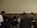 A group of people standing in a circle, appearing to be engaged in a moment of reflection or prayer. The setting is indoors with a neutral-colored background.