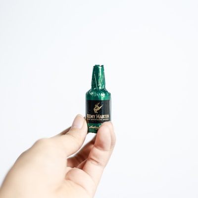 A hand holding a small chocolate in the shape of a miniature bottle, wrapped in green foil with a label that reads Rémy Martin Fine Champagne Cognac. The background is plain and white, highlighting the focus on the miniature chocolate bottle.
