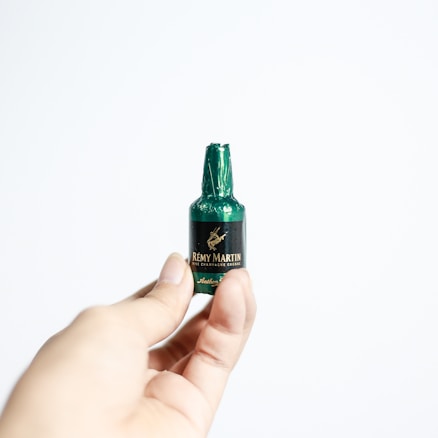 A hand holding a small chocolate in the shape of a miniature bottle, wrapped in green foil with a label that reads R&eacute;my Martin Fine Champagne Cognac. The background is plain and white, highlighting the focus on the miniature chocolate bottle.