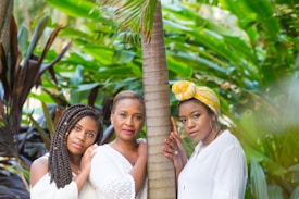 Three women stand closely together next to a palm tree, surrounded by lush green tropical foliage. They are wearing white clothing, and one woman has a yellow headwrap. Their expressions are serene and they appear to be enjoying the natural setting.