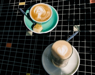 Two coffee drinks are served on a tiled table. One is in a turquoise cup with a saucer, accompanied by a piece of cake and a spoon resting on the side. The other is in a clear glass situated on a white saucer, both showcasing latte art with heart shapes on top.