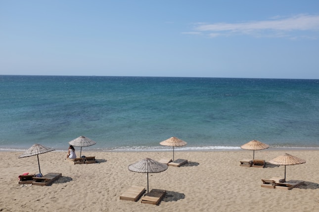 A serene beach scene with several straw umbrellas and wooden sun loungers arranged on the sandy shore. The calm blue sea extends into the horizon under a clear sky. A single person is sitting under one of the umbrellas, adding a sense of solitude and relaxation to the atmosphere.