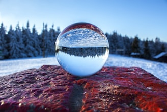 A crystal ball placed on a textured red rock reflects a snowy forest landscape. Tall evergreen trees in the background are covered in snow, creating a serene winter scene. The sky above is clear with a bright blue hue, enhancing the crispness of the image.
