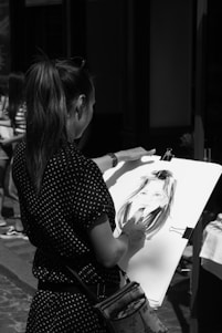 A person is drawing a portrait on a large sketchpad. The artwork features a face, and the artist holds a drawing tool. The setting appears to be outdoors with other people in the background. The image is in black and white, focusing on the artistry and technique.