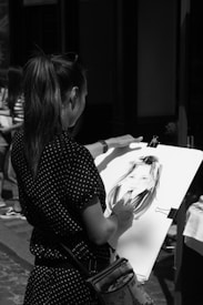 A person is drawing a portrait on a large sketchpad. The artwork features a face, and the artist holds a drawing tool. The setting appears to be outdoors with other people in the background. The image is in black and white, focusing on the artistry and technique.