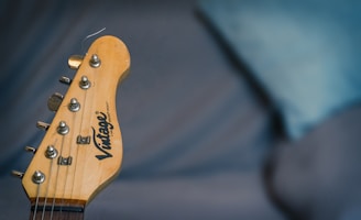 The image displays a close-up of a guitar headstock with tuning pegs and strings visible. The background is blurred, featuring a blue and gray setting.