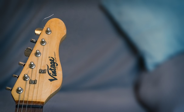 The image displays a close-up of a guitar headstock with tuning pegs and strings visible. The background is blurred, featuring a blue and gray setting.